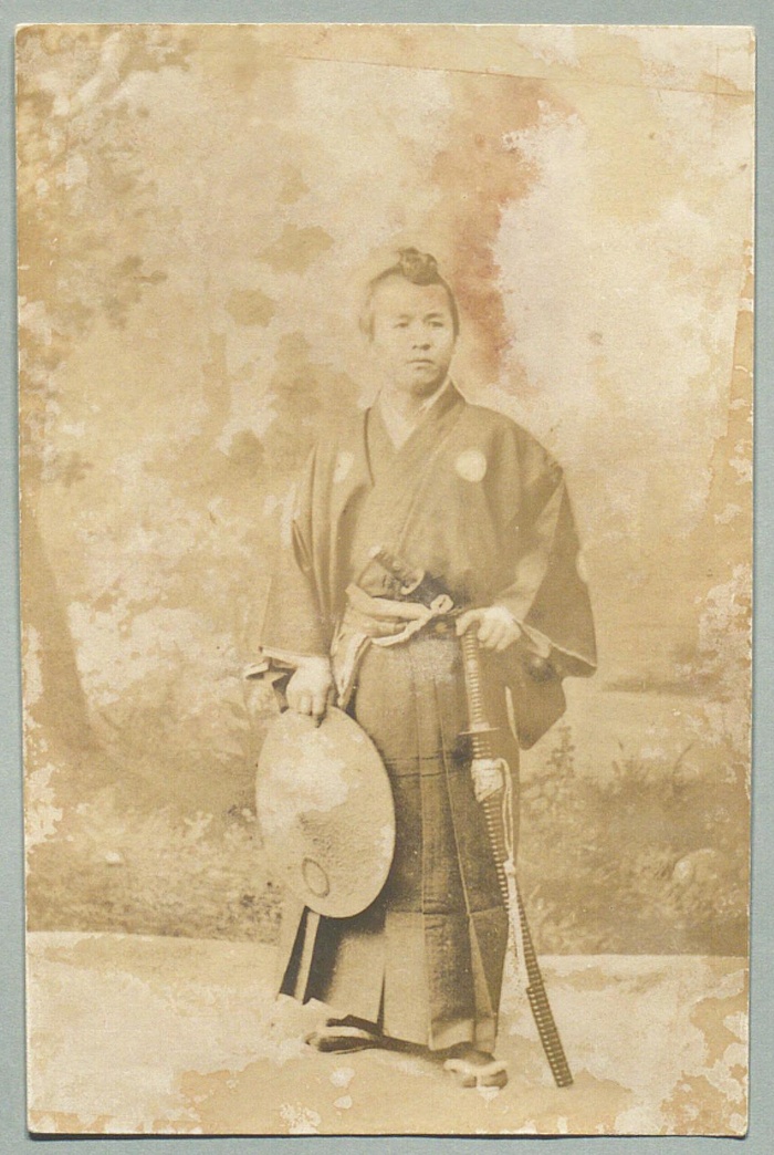 Faded photograph of a young man in kimono and hakama with two swords and a topknot, holding a round flat hat, in front of a painted backdrop