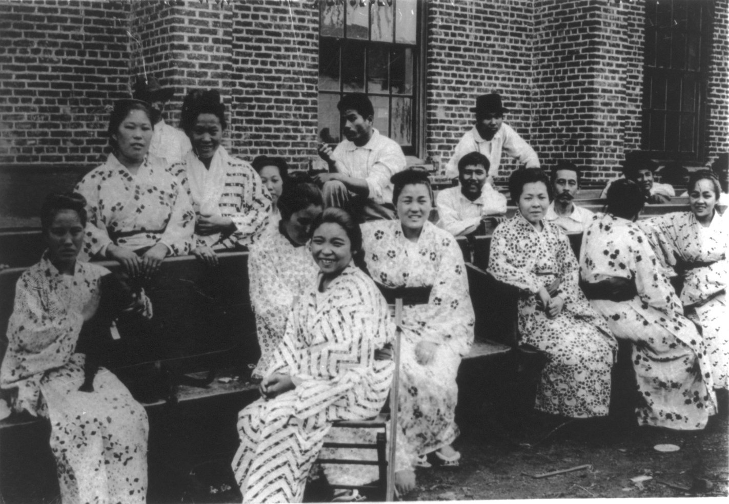 Young Japanese women in light patterned kimono, several smiling, sit on benches outside a brick building in Hawaii, with Japanese men behind them