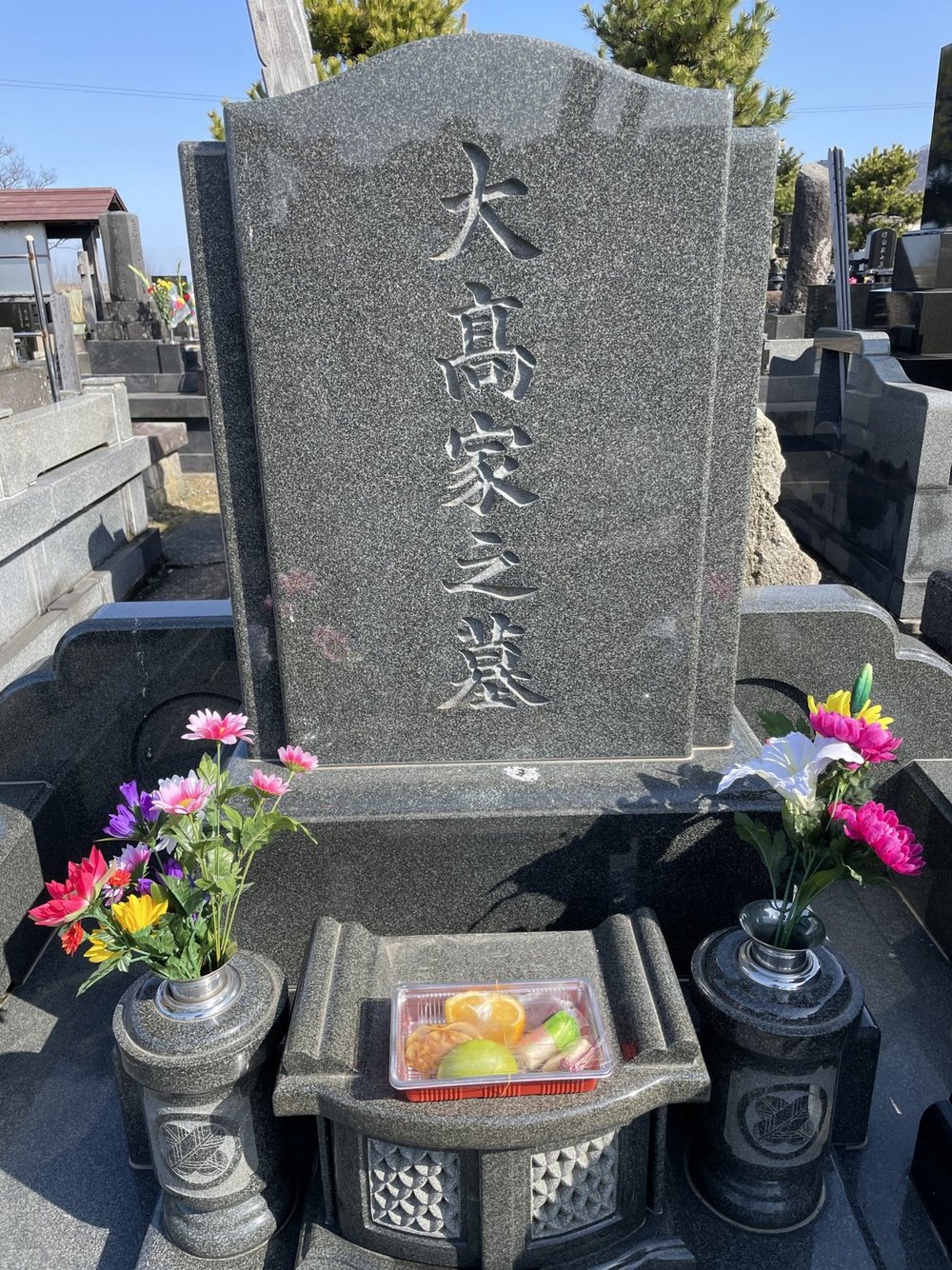 A dark granite family gravestone carved with the characters 大高家之墓, with bright flowers in vases on both sides and an offering of fruit in front