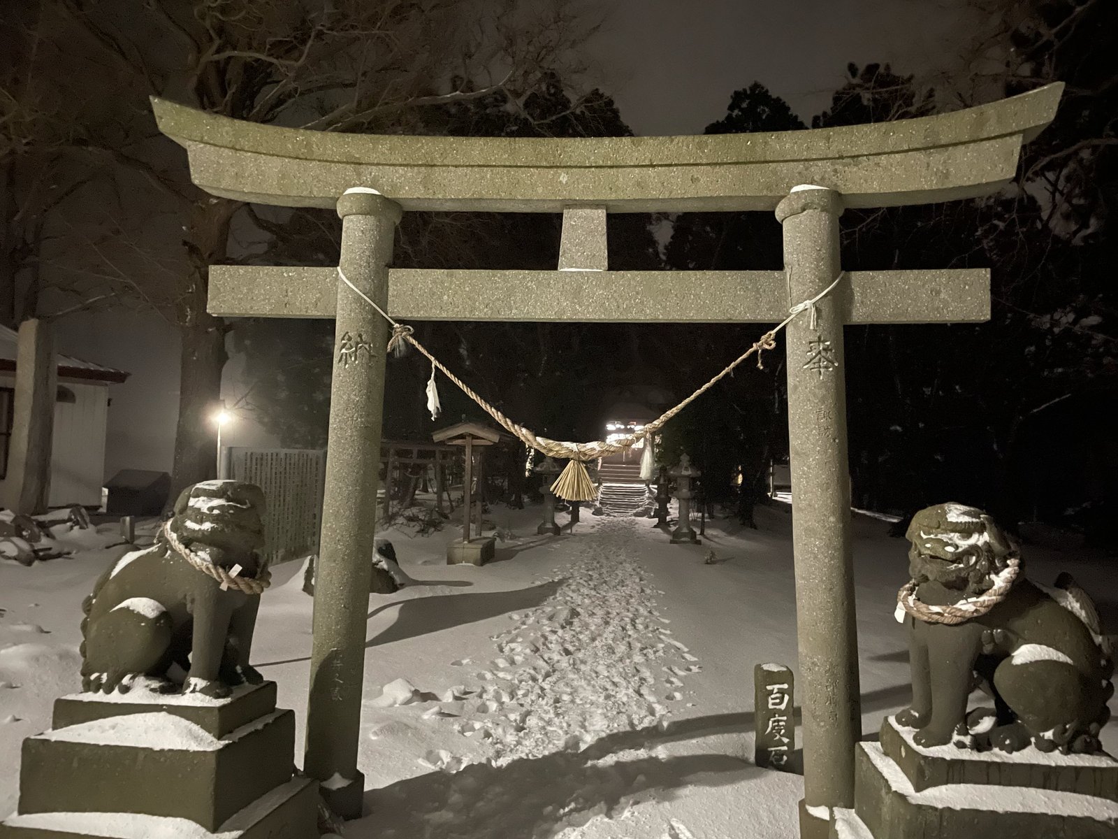 A stone torii gate hung with a thick straw rope, guarded by two stone lion-dogs, at the entrance of a shrine on a snowy night